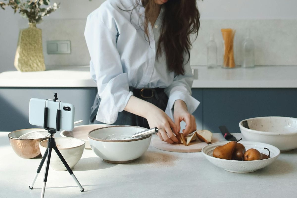 A woman preparing ingredients in a modern kitchen while recording with a smartphone on a tripod.