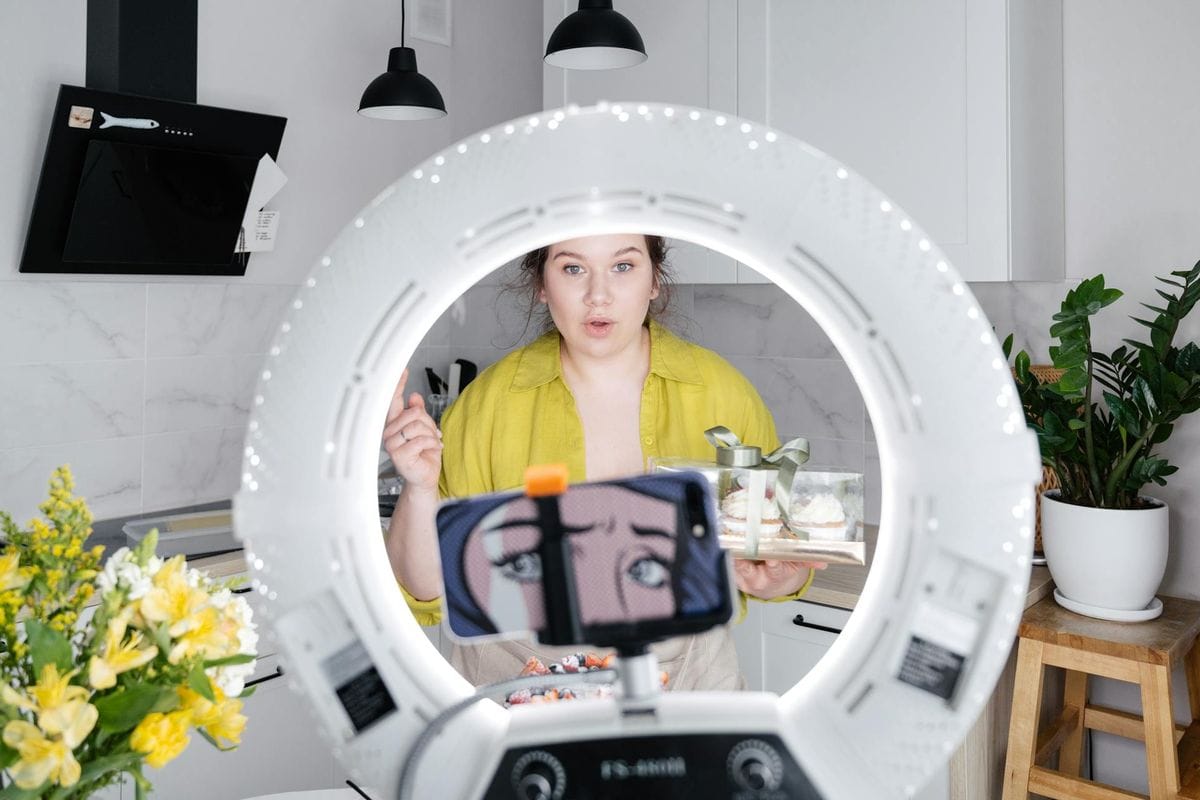 A young woman vlogs in a modern kitchen, using a smartphone and ring lamp.