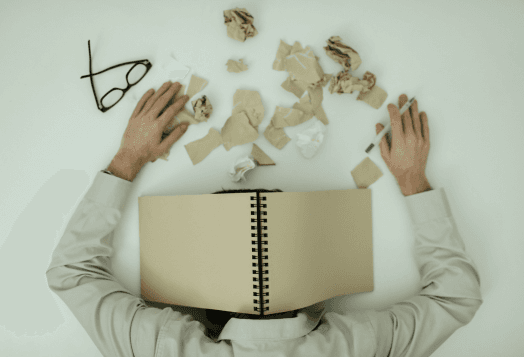 A man lying under a book with crumpled papers around his head, depicting stress