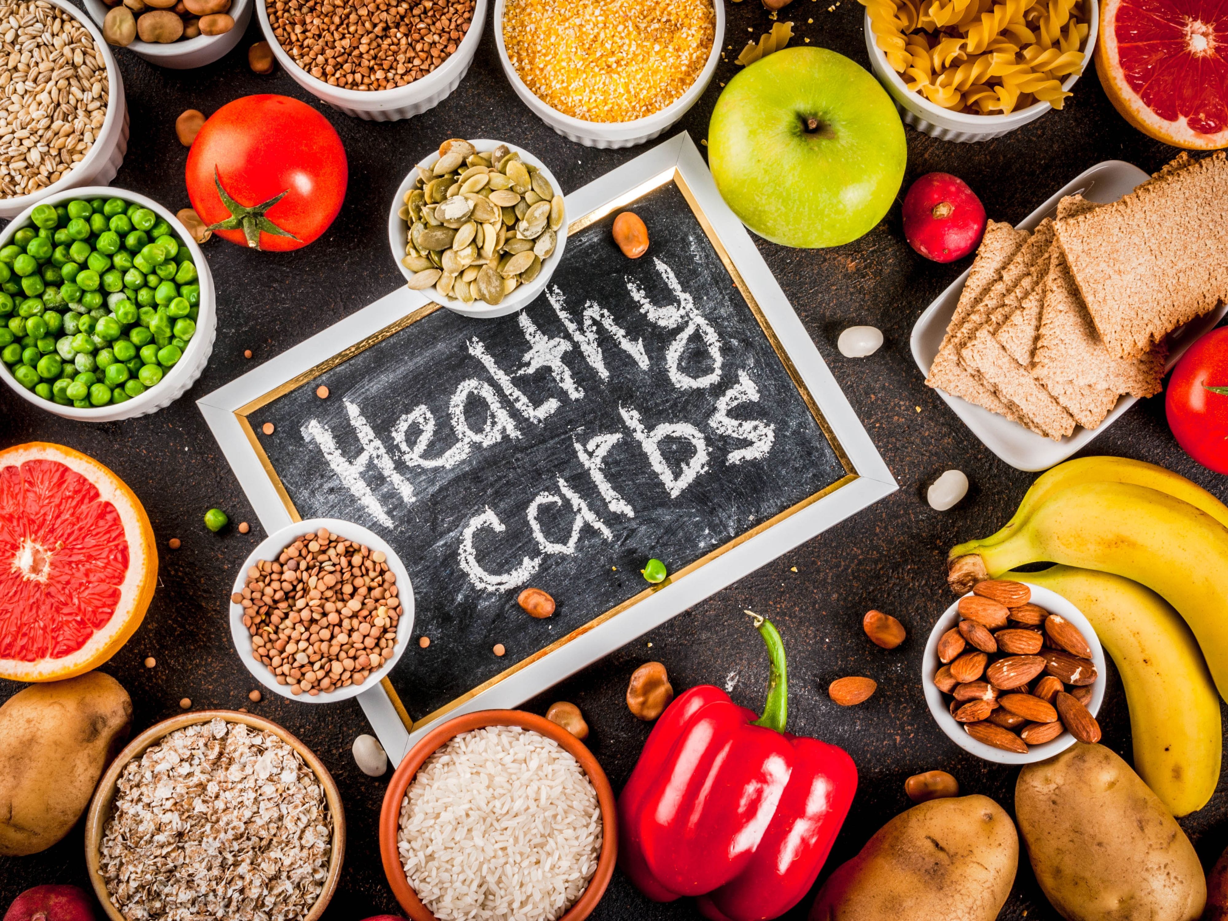 A chalkboard with "Healthy Carbs" written on it, surrounded by nutritious carbohydrate-rich foods such as fruits, vegetables, whole grains, nuts, and legumes