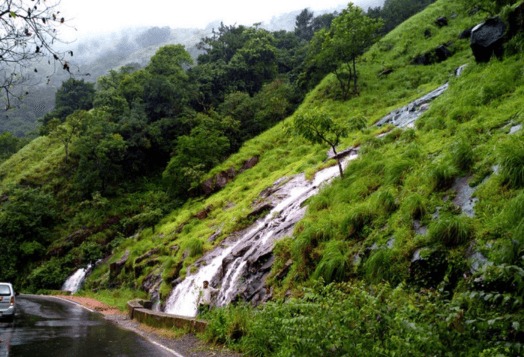 A landscape photo of a green, hilly area in Chikmagalur, Karnataka, with a waterfall cascading down the hillside and a road at the bottom