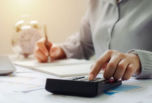 A person's hands using a calculator and writing in a notebook, with an alarm clock in the background