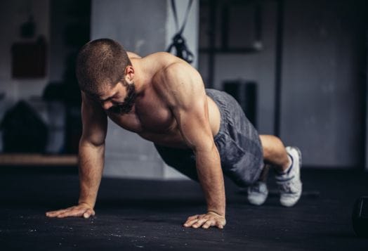 Man with beard doing a push-up on a dark gym floor indoors