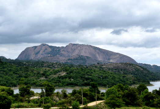 The massive granite monolith of Savandurga hill, with trees and a lake in the foreground under a cloudy sky