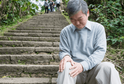 An elderly man with a pained expression sits on a stone step and holds his knee