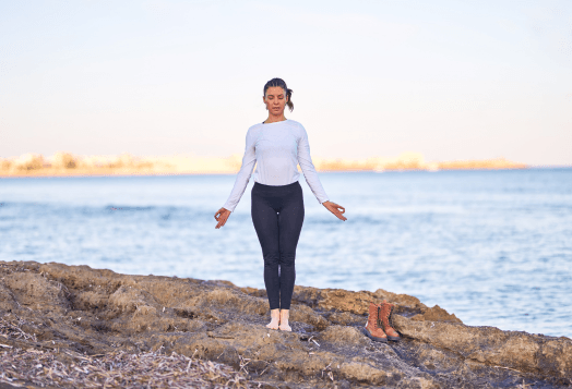 A woman demonstrates Mountain Pose (Tadasana) on a rocky shore by the sea