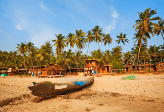 A sandy beach in Goa with a boat resting on the shore, surrounded by palm trees and beach huts under a blue sky