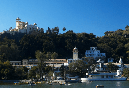 A scenic view of a large, red and white building on the banks of a misty river in Rishikesh, Uttarakhand, with lush green mountains in the background