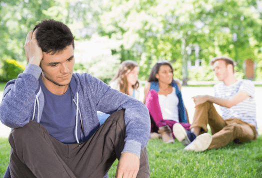 A young man sits alone in the foreground, looking sad, while a group of friends socializes in the background