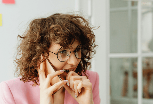 A woman with curly hair and glasses bites her nails while on the phone, looking anxious