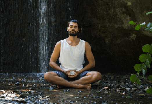 A man with a beard sits in a meditative pose with his eyes closed, with a dark, rocky background