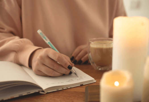 A person writes in a notebook, with a cup and lit candles nearby