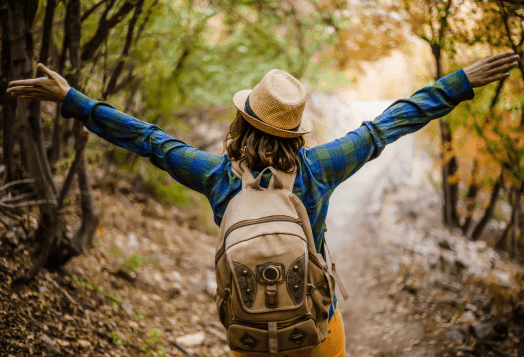 A person wearing a hat and backpack stands on a trail with their arms outstretched, facing away from the camera