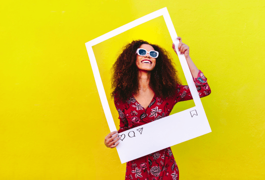 A smiling woman with curly hair and sunglasses holds up a large cutout of an Instagram post, framing herself