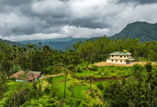 A landscape photo of a lush green valley in Coorg, Karnataka, with two houses and mountains under a cloudy sky