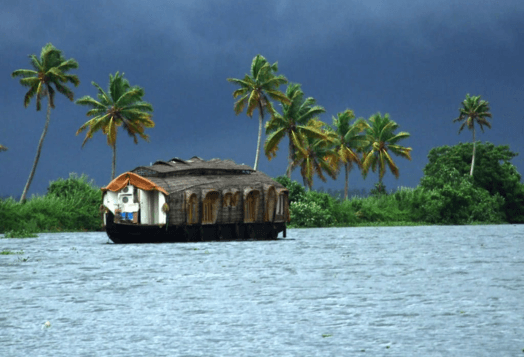 A houseboat floats on a backwater in Alleppey, Kerala, with palm trees and a cloudy sky in the background