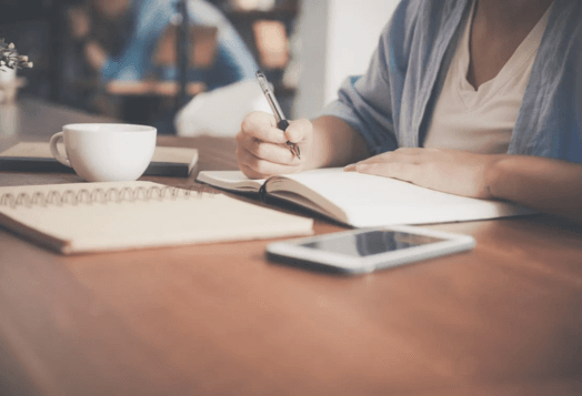 Person writing in a notebook on a wooden table with phone