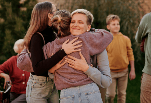 Two women embracing outdoors, a group of people nearby