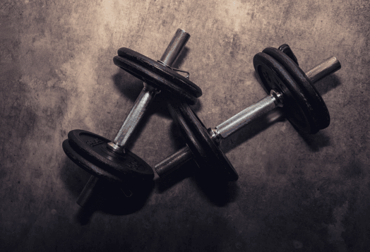 A top-down view of two small, adjustable dumbbells lying on a dark, textured floor