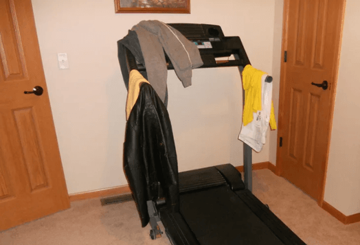 A treadmill in a room with light brown walls and carpet, with clothes draped over the handles