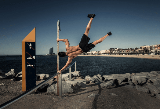 A muscular man performing a flag pole exercise on a horizontal bar at an outdoor gym, with a beach and the ocean in the background