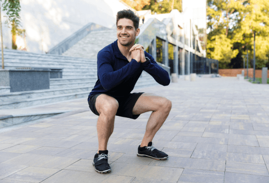 A smiling man in a long-sleeved navy shirt and black shorts doing a squat outdoors on a paved area