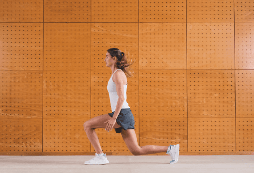 A woman in a white tank top and black shorts doing a lunge with her right leg forward against a wooden-paneled wall