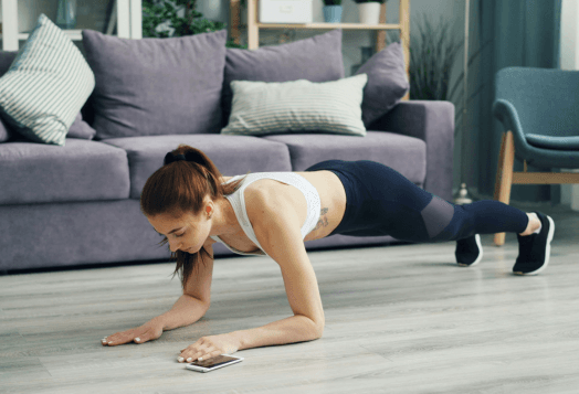 A woman in a sports bra and leggings doing a forearm plank in her living room, with her phone on the floor in front of her