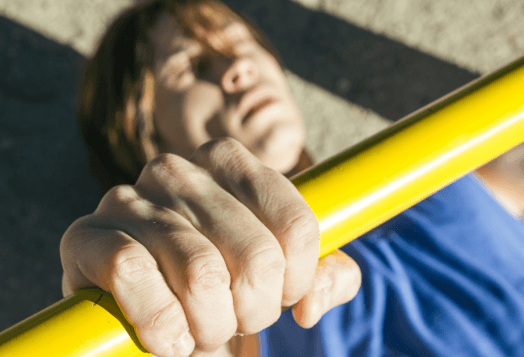 A close-up of a person's hand gripping a yellow bar, with the person's blurred, pained face in the background