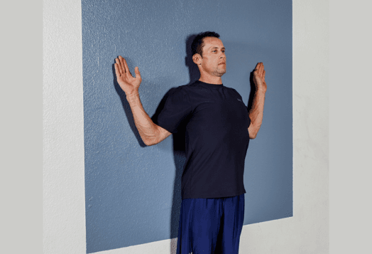 A man in a navy t-shirt and blue shorts is standing against a two-toned wall, performing a chest stretch with his arms bent at 90-degree angles and his hands pressed against the wall