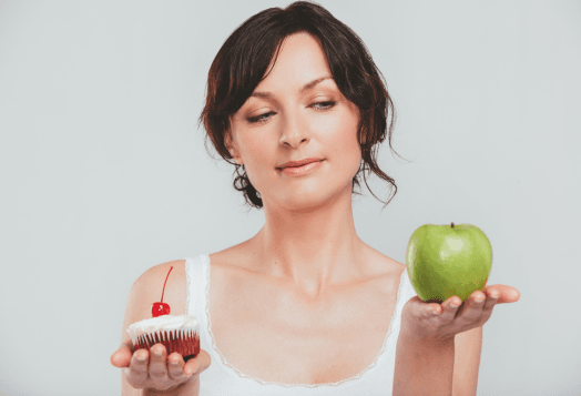 A woman holding a cupcake in one hand and an apple in the other, contemplating