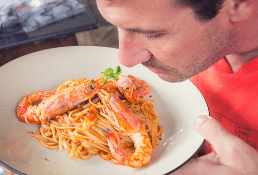 A man in a red shirt smelling a plate of pasta with prawns