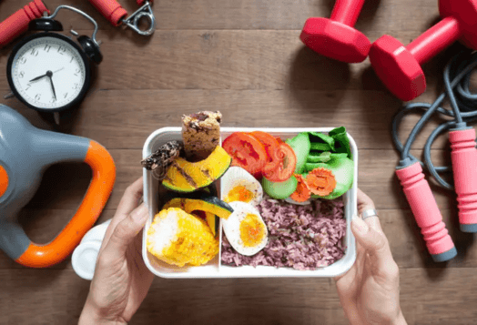 A top-down view of a person holding a bento box filled with healthy foods like rice, boiled eggs, and vegetables, surrounded by workout equipment such as dumbbells and a jumping rope