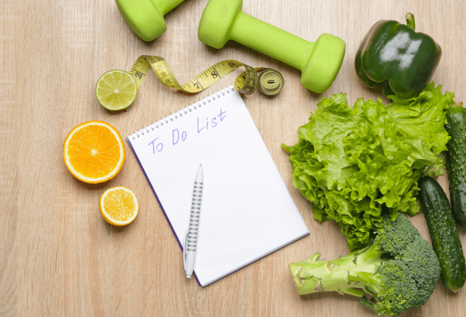 An overhead shot of a 'To Do List' notepad surrounded by various health-related items, including green vegetables, fruits, and light green dumbbells