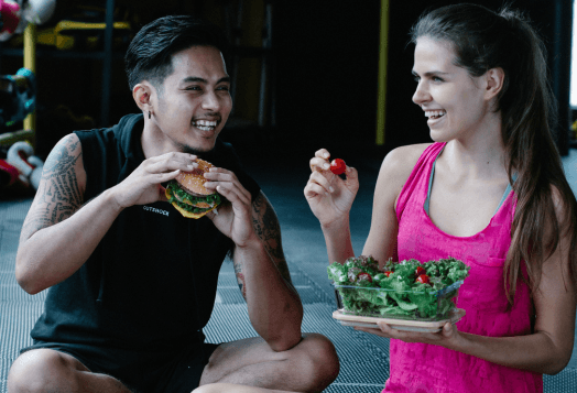 A man and a woman in workout clothes smiling and sitting on the floor after a workout, with the man eating a sandwich and the woman holding a container of salad