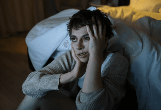 A tired-looking woman sitting on the floor, holding her head with a blanket on her lap