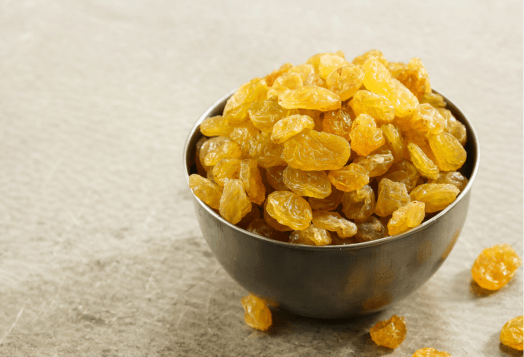 A close-up of a dark metal bowl filled with golden raisins, with some raisins scattered around it