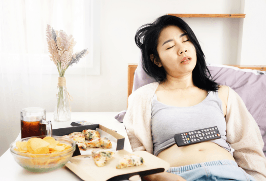 A woman asleep on a bed with a remote control on her stomach, surrounded by unhealthy food