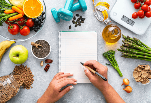 A top-down view of a person's hands writing in a spiral notebook, surrounded by fresh fruits, vegetables, seeds, dumbbells, and a digital scale