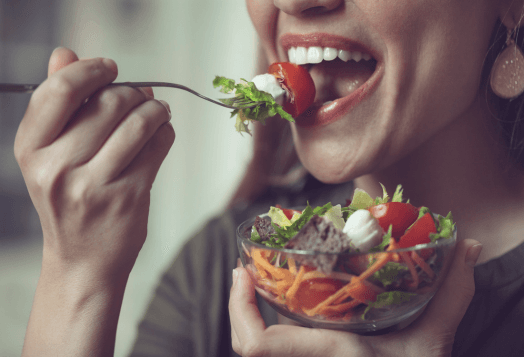 A close-up of a person's mouth and hand, taking a bite of a salad with a fork