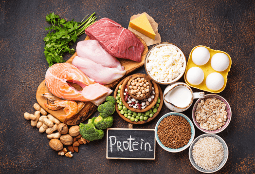 An overhead shot of a variety of foods rich in protein, including meat, fish, eggs, dairy, nuts, and legumes, with a small chalkboard sign that says 'Protein'
