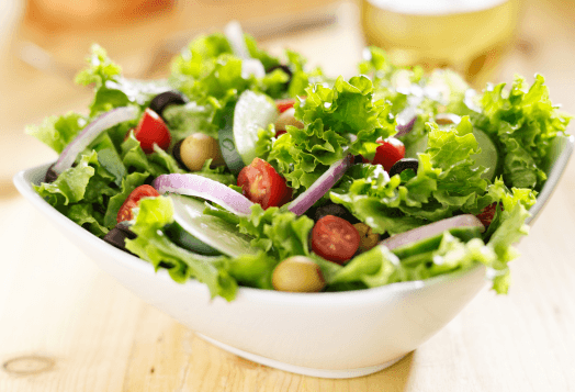 A close-up of a fresh green salad in a white bowl, with lettuce, tomatoes, onions, olives, and garbanzo beans