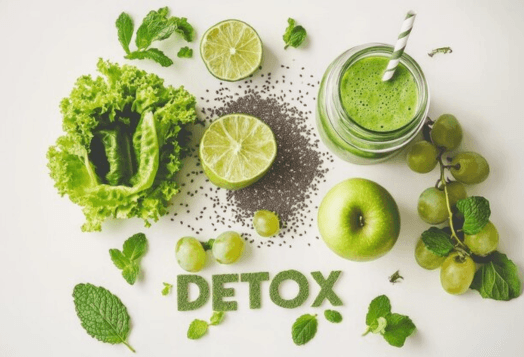 A top-down view of a green smoothie in a jar with a straw, surrounded by green fruits and vegetables like grapes, lettuce, lime, and mint leaves, with the word 'DETOX' spelled out in chia seeds and mint leaves