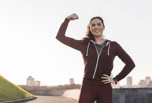 A smiling woman in workout clothes flexing her bicep outdoors