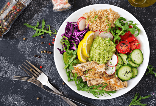 A top-down view of a plate with slices of grilled chicken, quinoa, and a salad with avocado, radishes, cabbage, and cucumbers