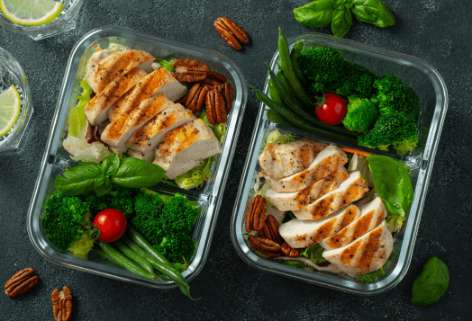 Two meal prep containers with sliced grilled chicken, broccoli, green beans, and pecans