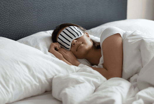 A woman in a striped sleep mask, lying in bed under a white comforter