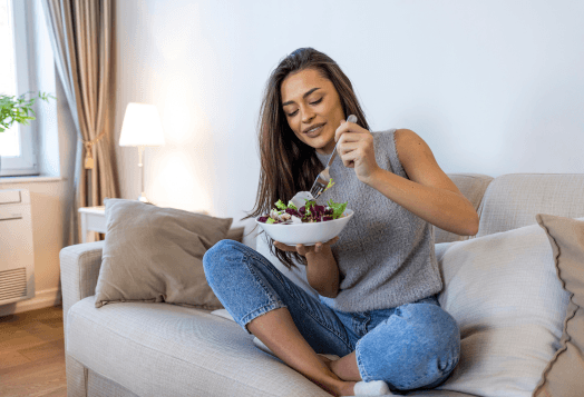A woman sitting cross-legged on a couch, smiling as she eats a salad from a white bowl