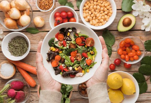 A person's hands holding a large bowl of salad surrounded by various fresh vegetables, seeds, and fruits on a wooden table