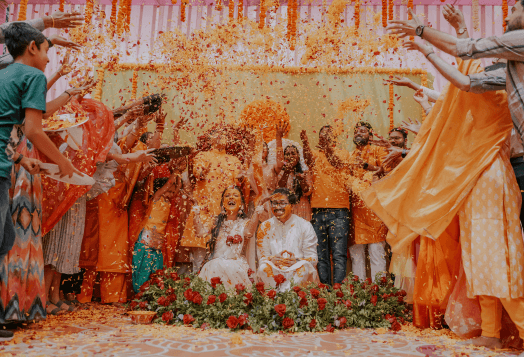 A newlywed couple sitting on a stage with a red floral arrangement, surrounded by people in festive clothes throwing flowers at them